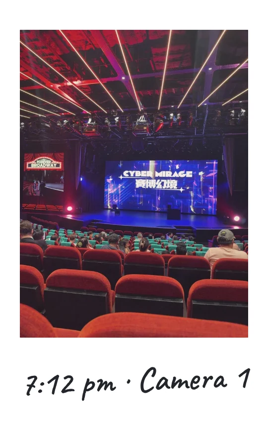 Camera 1, 7:12 pm: a theatre with a stage screen reading Cyber Mirage, the audience in red seats