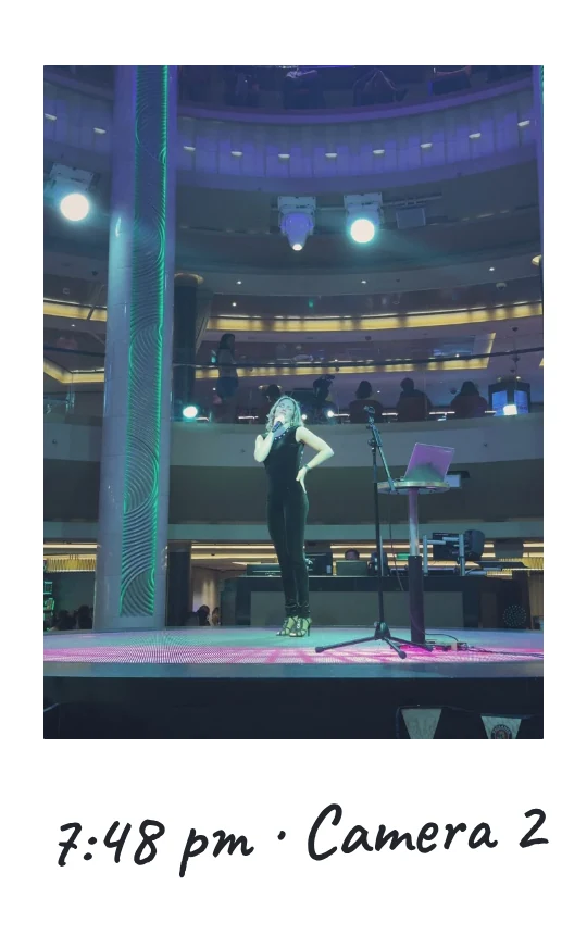 Camera 2, 7:48 pm: a singer in black on a stage in a tall atrium with balconies above